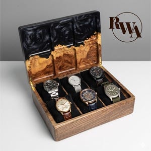May include: A wooden watch box with a black and brown resin top, containing seven watches with diverse band and face designs. The box is open, displaying the watches on black velvet cushions. The RWA logo is visible on the right.