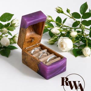 May include: A rectangular ring box crafted from natural burl wood and vibrant purple resin. The open lid reveals a beige velvet interior holding three silver-toned rings, including one with a central stone. The box is accented by white roses.