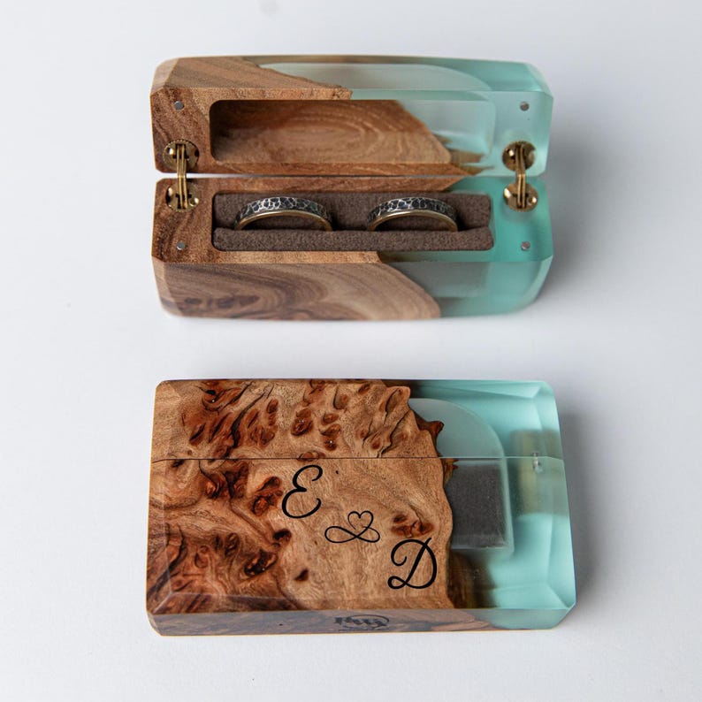 May include: A wooden ring box with a turquoise resin accent. The box is open, revealing two rings in a brown velvet interior. The lid is engraved with the letters "E & D" and a heart symbol. The box is made of wood and resin.
