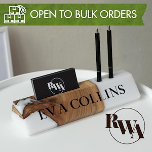 May include: A white and wood-tone business card holder with the name "EVA COLLINS" printed in black. The holder has a slot for cards and two pen holders. The text "OPEN TO BULK ORDERS" is displayed on a green banner.