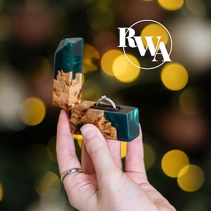 Può includere: Un cofanetto per anelli incernierato realizzato in radica di legno naturale e resina traslucida color verde acqua, contenente un anello color argento con una piccola pietra. La scatola presenta il logo bianco RWA in un cerchio. Il design unisce legno e resina.
