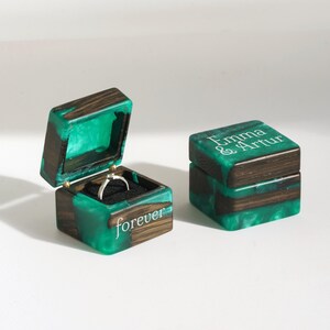 May include: Two square ring boxes made of dark wood and vibrant green resin. One box is open, revealing a silver ring inside on a black cushion, with the word forever printed on the side. The closed box features the text Emma & Artur on top.