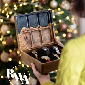 May include: A rectangular wooden watch box with a hinged lid featuring a black resin inlay and natural wood grain. The interior contains six black velvet cushions, with five wristwatches displayed inside. The logo RWA is visible.