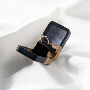 Op de afbeelding: Een kleine, rechthoekige ringdoos met een combinatie van natuurlijke houtnerf en gladde zwarte hars. De binnenkant is bekleed met zwart fluweel en bevat een zilverkleurige ring met een kleine, heldere steen in het midden van de ringband.