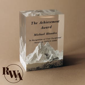 May include: A rectangular clear resin award block featuring a white mountain range sculpture embedded at the base. The front surface displays black text reading The Achievement Award, Michael Rhoades, In Recognition of Your Exceptional Success at NEXUS CORE.