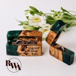 Può includere: Due scatole rettangolari per anelli realizzate in radica di legno naturale e resina verde scuro traslucida. La scatola più grande contiene due anelli e presenta il testo inciso "My heart belongs to you". La scatola più piccola è aperta e contiene un anello.