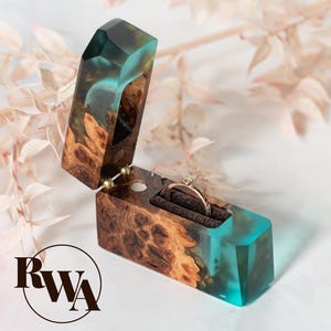 May include: A handcrafted ring box made of wood and teal resin. The box is open, revealing a gold ring with a diamond. The box has a unique wood grain pattern and is set against a soft, neutral background. The RWA logo is in the lower left corner.