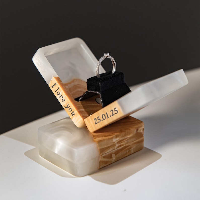 Op de afbeelding: Een rechthoekig ringdoosje van witte hars en lichtbruin hout, met een zwart fluwelen inzetstuk voor een zilveren ring. Het doosje staat open en toont de tekst "I love you" aan de linkerkant en de datum "25.01.25" aan de rechterkant.