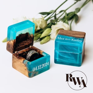 May include: Two square ring boxes made of natural wood and translucent turquoise resin. One box is open, revealing a silver patterned ring inside, with the date 04.12.2024 engraved on the side. The closed box features the names Alex and Kailey.