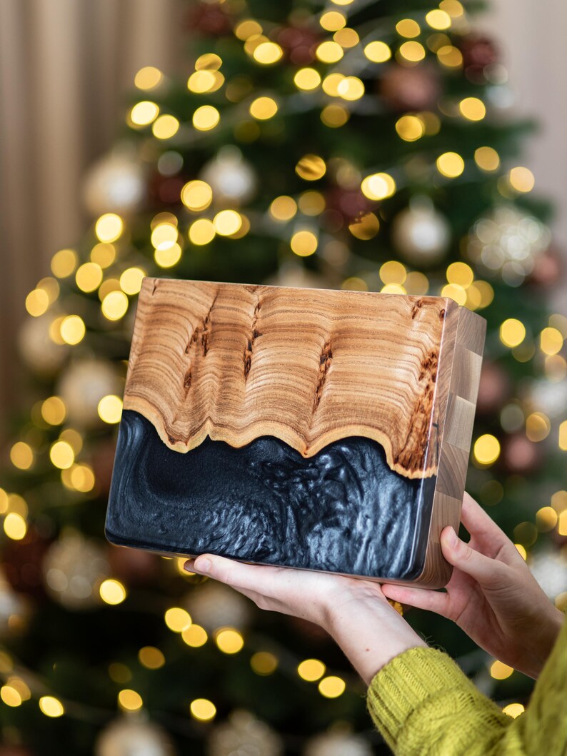 May include: A rectangular wooden serving board featuring a natural light brown wood grain top section with a wavy edge and a solid, opaque black resin bottom section with a smooth, glossy finish and a thick, block-style side profile.