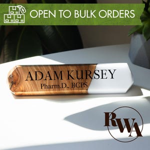 May include: A wooden desk nameplate with the name "ADAM KURSEY" and credentials "Pharm.D., BCPS" in black text. The nameplate has a wood grain finish on one end and a white, angled finish on the other. The text "OPEN TO BULK ORDERS" is in a green banner.