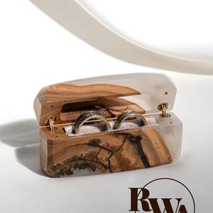 Könnte beinhalten: Eine rechteckige Ringbox aus Naturholz und durchscheinendem weißem Harz mit Klappdeckel und weichem Innenfutter, das zwei metallische Ringe mit strukturierter Oberfläche enthält. Das Logo RWA ist unten rechts im Bild zu sehen.