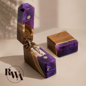 Può includere: Un portaanelli rettangolare in legno naturale e resina marmorizzata viola, con coperchio incernierato a chiusura magnetica e un inserto in velluto beige che ospita un anello argentato. Una seconda scatola chiusa abbinata è accanto. Logo RWA.