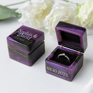 May include: Two square ring boxes made of dark wood and purple resin. One box is closed with the text Sophia & David engraved on top. The other box is open, revealing a silver ring with a small stone inside, and the date 03.10.2023 on the front.