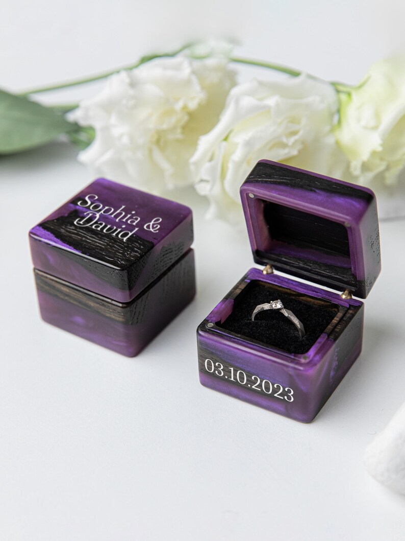 May include: Two square ring boxes made of dark wood and purple resin. One box is closed with the text Sophia & David engraved on top. The other box is open, revealing a silver ring with a small stone inside, and the date 03.10.2023 on the front.
