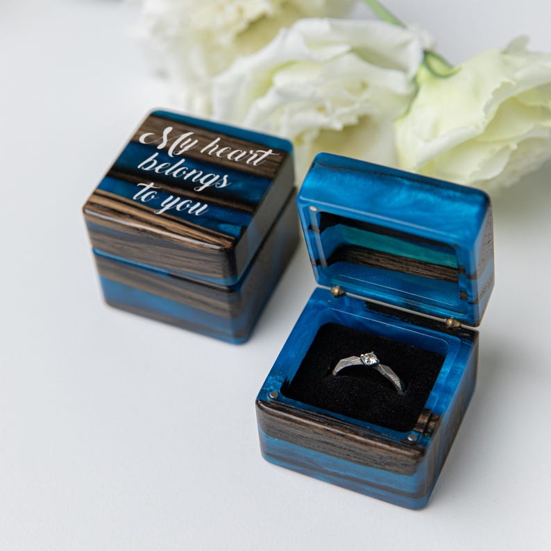 May include: Two square ring boxes made of dark wood and vibrant blue resin. One box is closed, featuring the white script text "My heart belongs to you" on the lid. The other box is open, revealing a black velvet interior holding a silver ring.