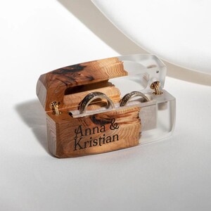 May include: A wooden ring box with a clear acrylic lid, holding two rings. The box is engraved with the names "Anna & Kristian". The rings are silver with dark accents. The box is open, revealing the rings.