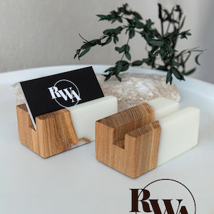 May include: Two rectangular business card holders made of natural wood and opaque white resin. One holder contains a black business card with the white circular logo RWA. The wood features a visible grain pattern and a straight vertical slot.