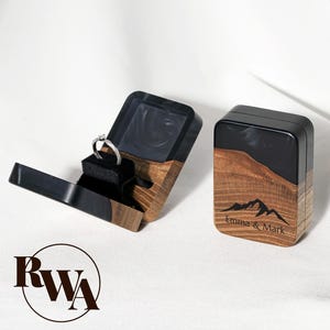 May include: A wooden ring box with a black top and a mountain design, open to reveal a silver ring on a black velvet cushion. The box is engraved with the names "Emma & Mark". The box is made of wood and black resin.