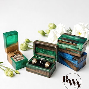 May include: Wooden ring boxes with green and blue resin accents. The boxes are open, displaying rings. The boxes are rectangular with a hinged lid. The background is white with white flowers.