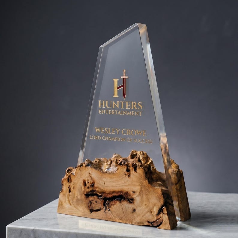 May include: A clear acrylic award with a wooden base. The award features the text "Hunters Entertainment" and "Wesley Crowe, Lord Champion of Success" in gold lettering. The base is a natural wood with a unique grain.