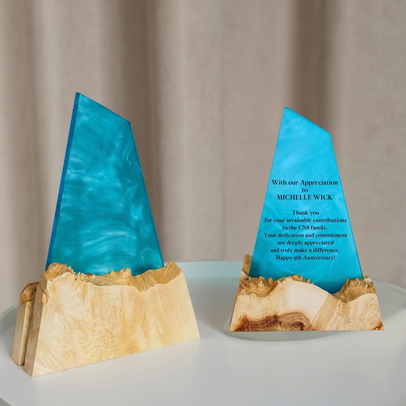 May include: Two decorative awards. Each award features a teal-colored, triangular resin top with a swirling pattern, mounted on a light-colored, textured wooden base. One award has text that reads "With our Appreciation to MICHELLE WICK".