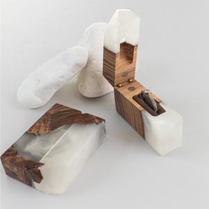 May include: Wooden ring box with a white resin base and a hinged lid. The box is open, revealing a ring on a brown velvet cushion. Two white stones are in the background. The box is approximately 3 inches long.