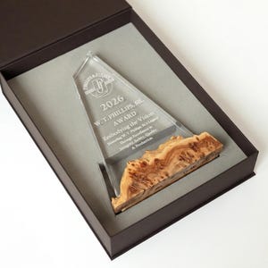May include: A clear acrylic award with a wooden base, presented in a dark brown box. The award is engraved with text including "2026 W.T. PHILLIPS, SR. AWARD" and "Embodying the Vision". The wooden base has a unique, natural grain pattern.