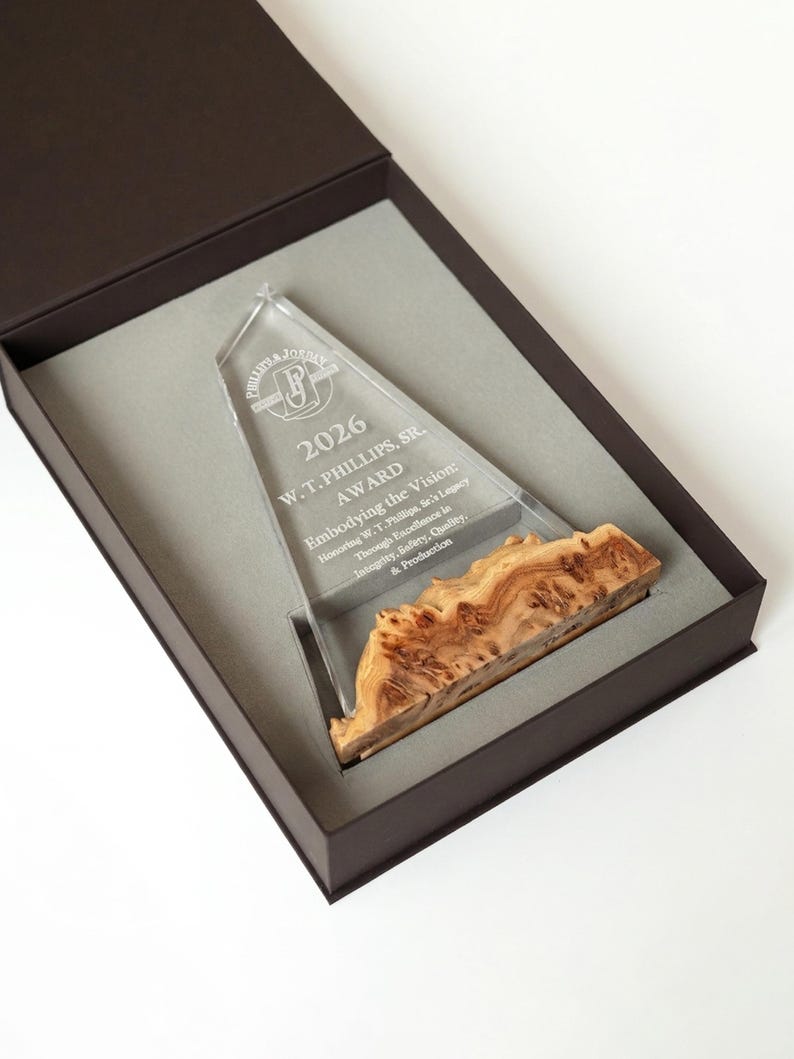 May include: A clear acrylic award with a wooden base, presented in a dark brown box. The award is engraved with text including "2026 W.T. PHILLIPS, SR. AWARD" and "Embodying the Vision". The wooden base has a unique, natural grain pattern.