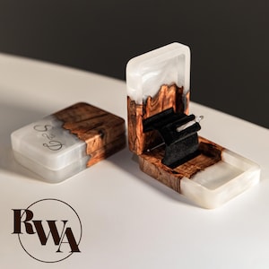 May include: Open wooden ring box with a white resin interior and a black velvet ring holder. The box is made of wood with a natural grain pattern and a white resin top. A second closed box is visible.