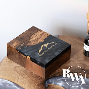 May include: A rectangular wooden box featuring a natural burl wood grain section and a smooth black resin inlay on the lid. The lid is engraved with a gold mountain graphic and the text "Sebastian & Rebecca" in a clean, sans-serif font.
