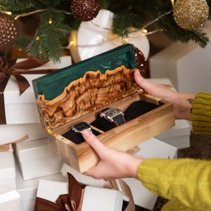 May include: A rectangular wooden watch box with a hinged lid featuring a live edge wood grain and a translucent emerald green resin inlay. The interior is lined with black fabric and contains two wristwatches held in place.