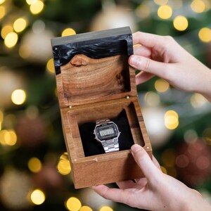 May include: A silver digital wristwatch with a rectangular face and metal link band rests inside a square wooden box. The box features a natural wood grain base and a dark, polished resin lid with a live edge detail, lined with black fabric.