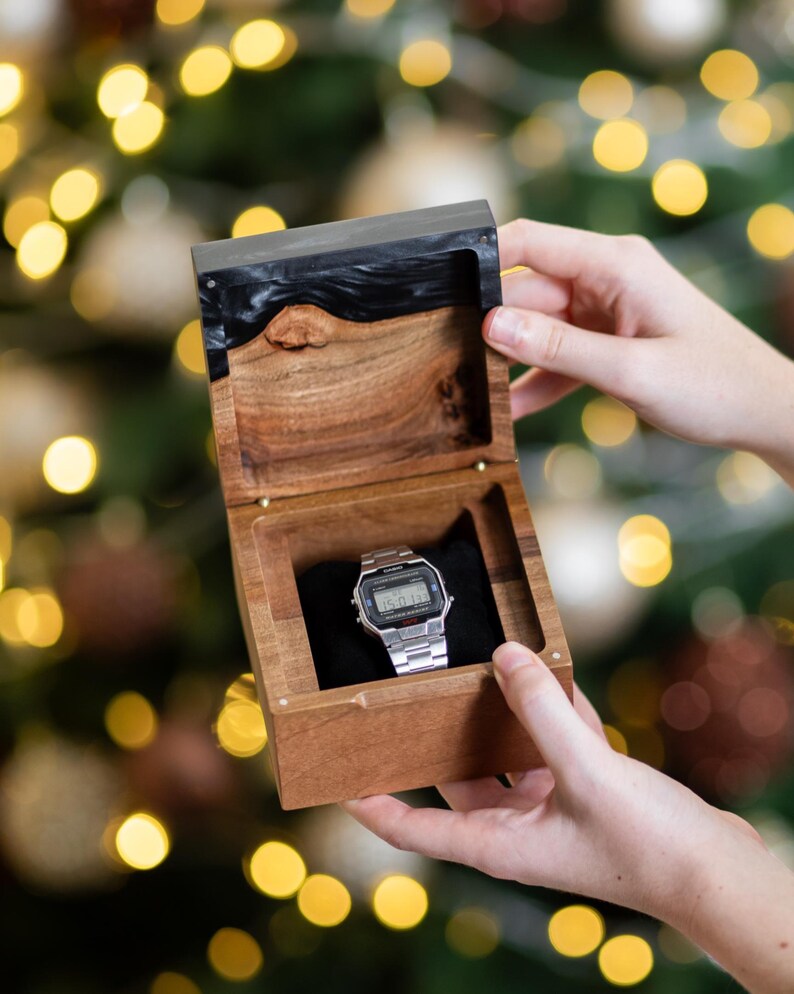 May include: A silver digital wristwatch with a rectangular face and metal link band rests inside a square wooden box. The box features a natural wood grain base and a dark, polished resin lid with a live edge detail, lined with black fabric.