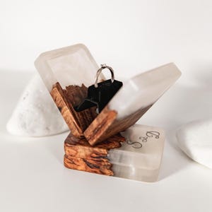 Op de afbeelding: Een vierkante ringdoos gemaakt van natuurlijk hout en doorschijnende witte hars. De doos staat open en onthult een zwarte inzet die een zilveren ring met een kleine steen vasthoudt. De buitenkant van de basis bevat de gegraveerde letters S en D.