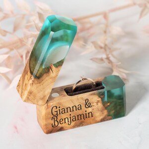 May include: A wooden ring box with a turquoise and clear resin top and interior. The box is open, revealing a gold ring with a small diamond. The names "Gianna & Benjamin" are engraved on the front.