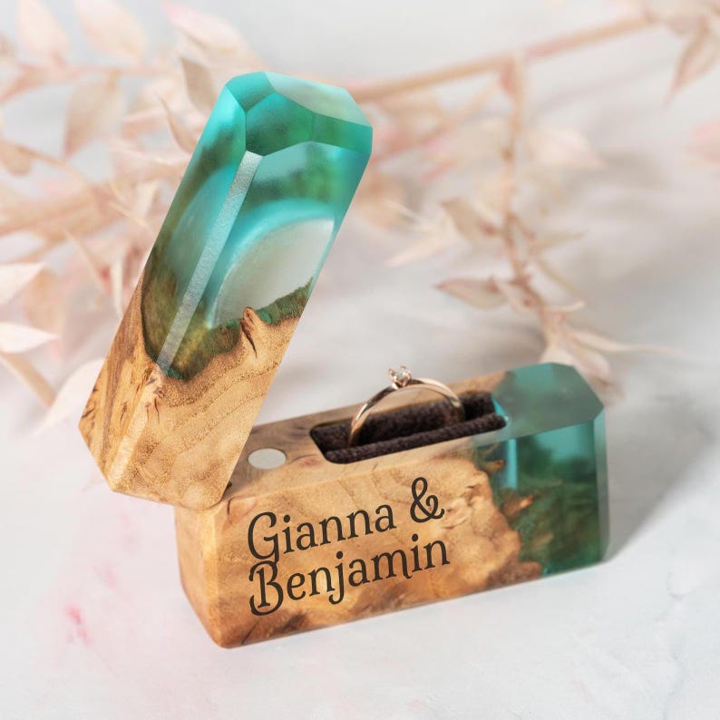 May include: A wooden ring box with a turquoise and clear resin top and interior. The box is open, revealing a gold ring with a small diamond. The names "Gianna & Benjamin" are engraved on the front.
