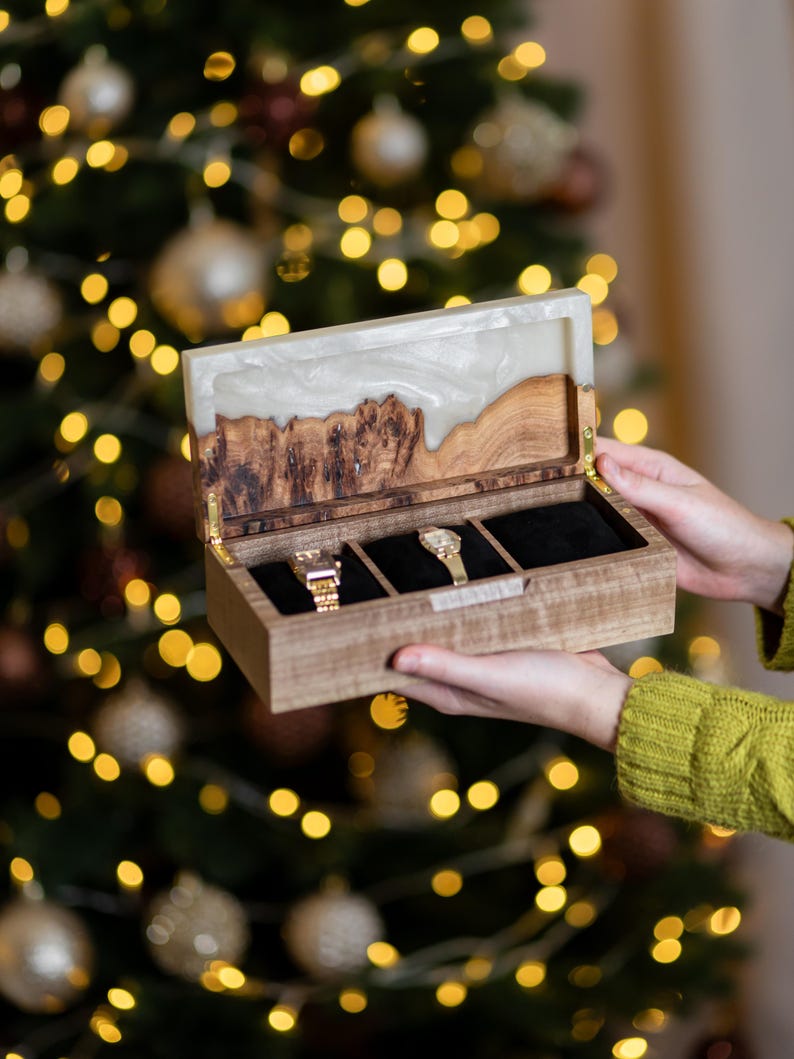 May include: A rectangular wooden watch box with a hinged lid featuring a white resin inlay. The interior is divided into three compartments lined with black fabric, containing two gold-toned wristwatches. The box has a natural wood grain finish.