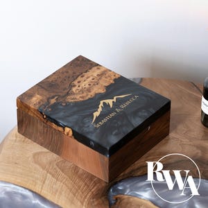 May include: A rectangular wooden box with a lid featuring a combination of natural burl wood and black resin. The lid is engraved with a gold mountain graphic and the text Sebastian & Rebecca. The box sides are made of solid brown wood.