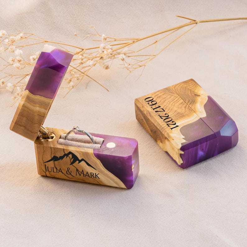 May include: A wooden ring box with a purple resin accent, open to reveal a silver ring. The box is engraved with "Julia & Mark" and a mountain range. The lid of the box next to it is engraved with the date "09.17.2021".