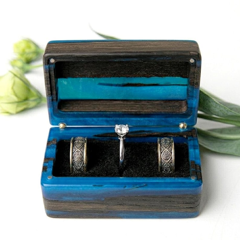 May include: A rectangular ring box crafted from dark wood and vibrant blue resin. The open box features a black velvet interior holding two patterned metal bands and a central silver ring with a single clear stone set in a prong mounting.