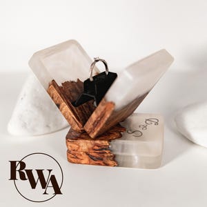 May include: A handcrafted ring box with a hinged lid, featuring a clear resin exterior with wood and a silver ring. The box is open, revealing the ring on a black holder. The base of the box has engraved initials. The box is on a white surface.