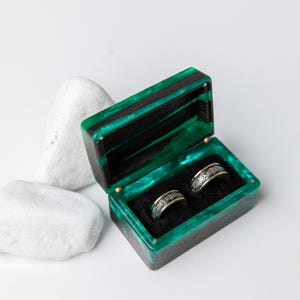 May include: A rectangular ring box made of dark wood and translucent emerald green resin with a hinged lid. The interior is lined with black fabric and holds two silver-toned metal rings featuring an intricate, repeating engraved pattern.