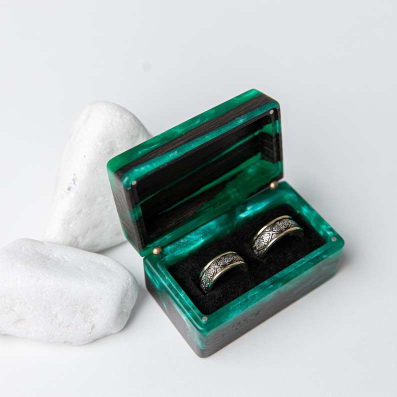 May include: A rectangular ring box made of dark wood and translucent emerald green resin with a hinged lid. The interior is lined with black fabric and holds two silver-toned metal rings featuring an intricate, repeating engraved pattern.