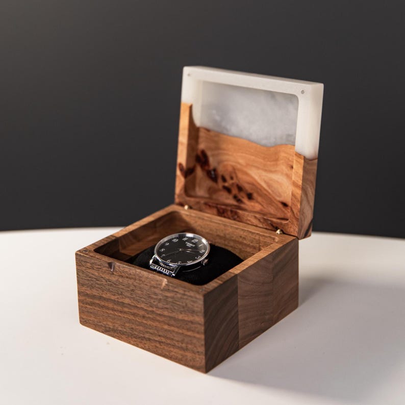 May include: A square wooden watch box with a hinged lid featuring a white resin top section. The box is crafted from dark brown wood with visible grain. Inside, a round wristwatch with a black face and silver hands rests on a black cushion.