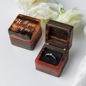 May include: Two square ring boxes made of dark wood and amber-colored resin. One box is closed with the white text Will you merry me? engraved on the lid. The other box is open, revealing a black velvet interior holding a silver ring with a stone.