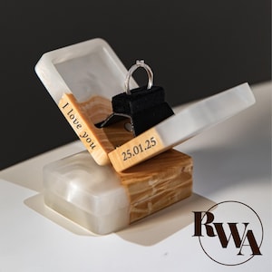 Peut inclure: Une bague de fiançailles en argent présentée dans un écrin en bois ouvert. La boîte est faite de bois et de résine blanche, avec les inscriptions "I love you" et "25.01.25". La bague repose sur un coussin en velours noir.