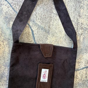 May include: A dark brown shoulder bag with a long strap. The bag has a flap closure and a rectangular patch with a logo. The bag is made of a textured material, and the strap has a lighter-colored lining.