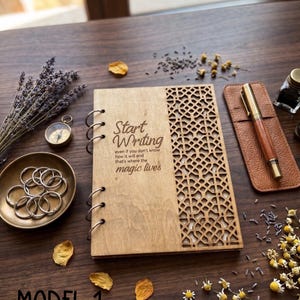 May include: A wooden binder with a laser-cut geometric pattern on the right side and the text "Start Writing even if you don't know how it will end that's where the magic lives" on the left. It features metal rings and is surrounded by dried flowers.