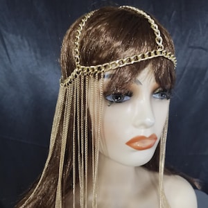 May include: A gold-colored chain headpiece with multiple strands of chain cascading down. The headpiece features a chain that goes around the head and a chain that goes over the top of the head. The headpiece is displayed on a mannequin.